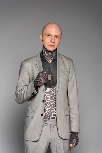 Schöner glatzköpfiger Mann mit Tätowierungen auf der Brust, der Anzug trägt und isoliert auf grau in die Kamera schaut — Stockfoto