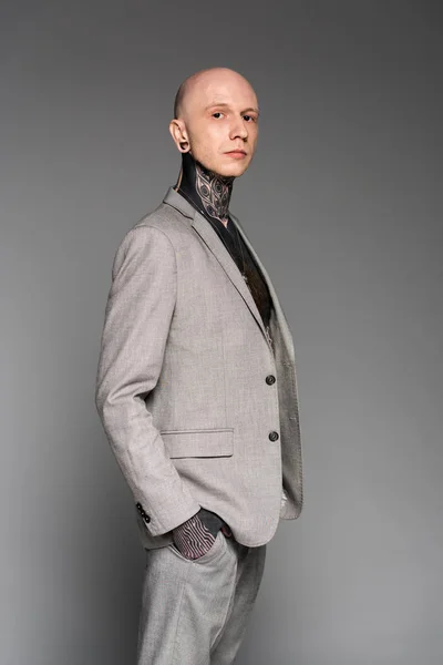 Seitenansicht eines gut aussehenden, tätowierten Mannes mit Glatze im Anzug, der mit der Hand in der Tasche steht und isoliert auf grau blickt — Stockfoto