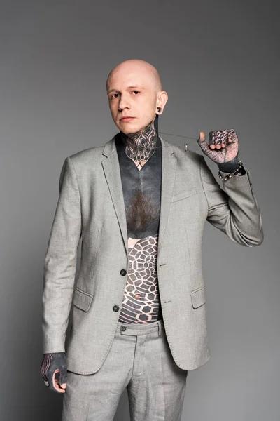 Стильный татуированный мужчина в сером костюме, держащий цепь и смотрящий на камеру, изолированную на сером — стоковое фото