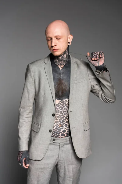 Hombre tatuado con estilo en traje gris que sostiene la cadena y mirando hacia abajo aislado en gris - foto de stock