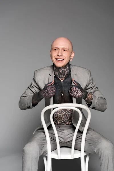 Hombre tatuado sonriente ajustando chaqueta de traje y sentado en silla en gris - foto de stock
