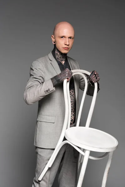 Confiado hombre calvo tatuado en traje sosteniendo silla blanca y mirando a la cámara aislada en gris - foto de stock