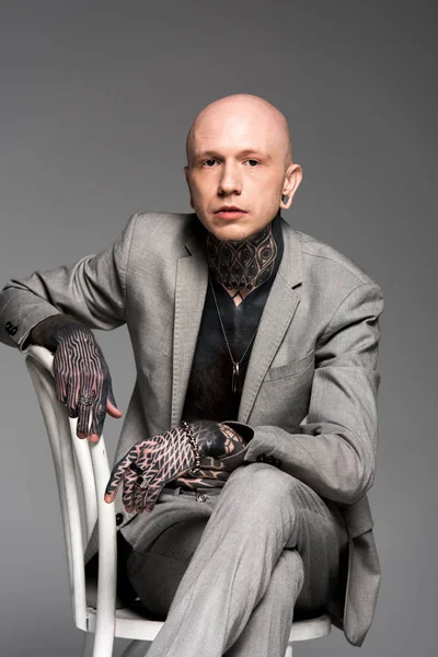 Уверенный лысый татуированный мужчина в костюме сидит на стуле и смотрит на камеру, изолированную на сером — стоковое фото
