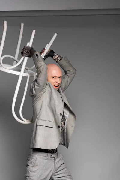 Aggressive tattooed man in suit holding chair above head and looking at camera on grey — Stock Photo