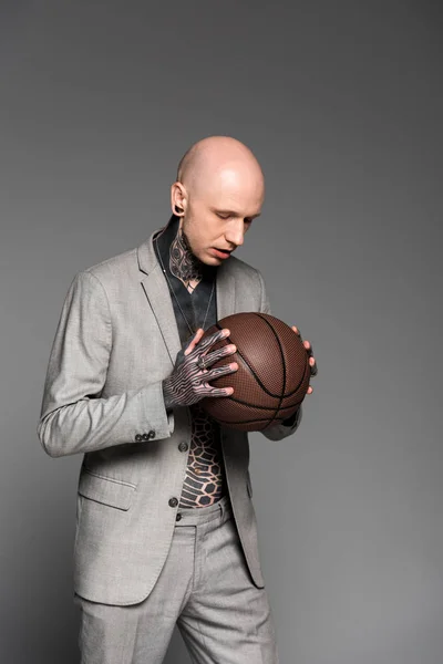 Серйозний лисий татуйований чоловік у костюмі тримає баскетбол ізольований на сірому — стокове фото