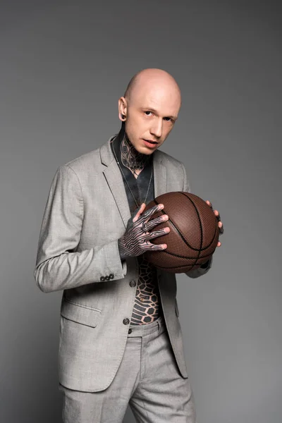 Guapo calvo tatuado hombre de traje sosteniendo pelota de baloncesto y mirando a la cámara aislada en gris - foto de stock