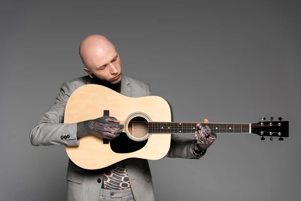 Красивый лысый татуированный мужчина в костюме играет на акустической гитаре, изолированной на сером — стоковое фото