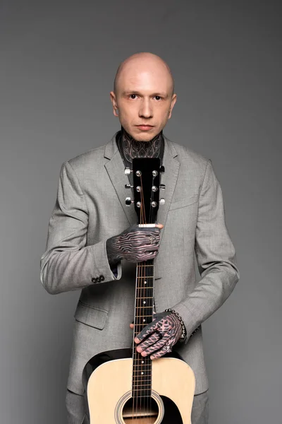 Handsome bald tattooed man in suit holding acoustic guitar and looking at camera isolated on grey — Stock Photo