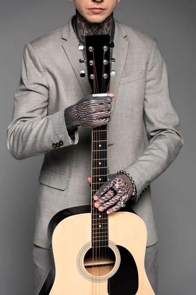 Recortado disparo de hombre tatuado en traje sosteniendo guitarra acústica aislado en gris - foto de stock