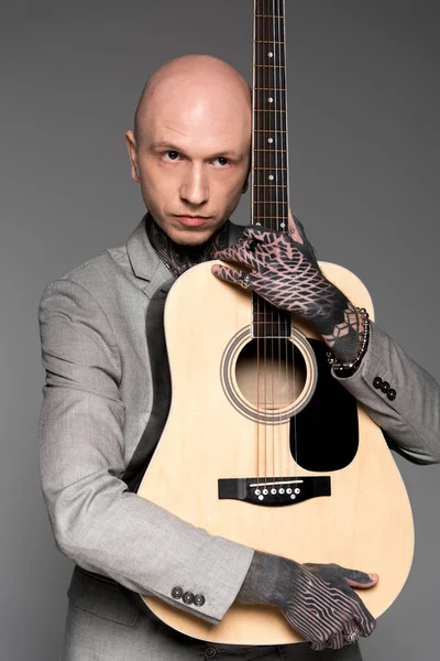 Guapo calvo tatuado hombre en traje abrazando guitarra acústica aislado en gris - foto de stock