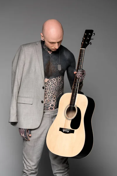 Hombre tatuado en el pecho desnudo con chaqueta de traje en el hombro mirando la guitarra acústica en gris - foto de stock