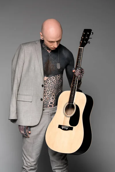 Bare-chested tattooed man with suit jacket on shoulder looking at acoustic guitar on grey — Stock Photo