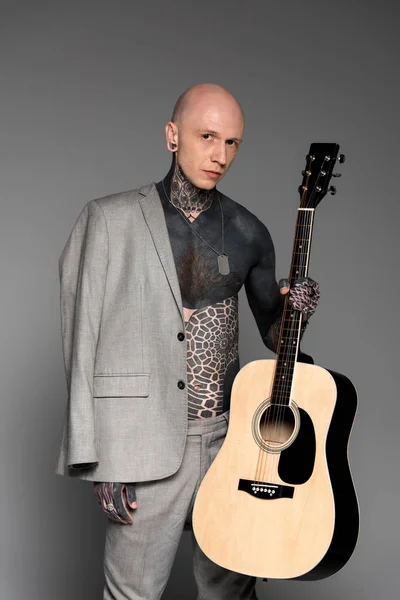 Bare-chested tattooed man with suit jacket on shoulder holding acoustic guitar and looking at camera on grey — Stock Photo