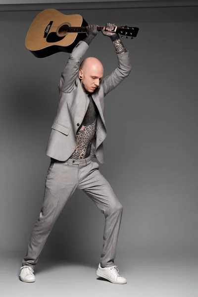 Повнометражний вигляд агресивного лисого татуйованого чоловіка в костюмі, що вражає акустичну гітару на сірому — стокове фото