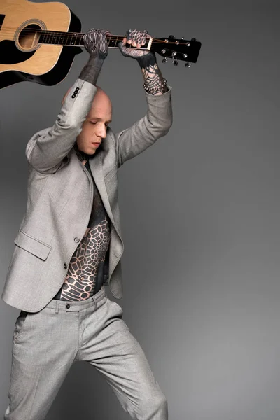 Aggressive bald tattooed man in suit hitting acoustic guitar on grey — Stock Photo