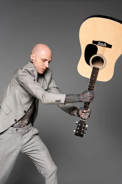 Angry tattooed man in suit hitting acoustic guitar isolated on grey — Stock Photo