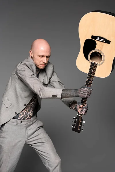 Aggressiver tätowierter Mann im Anzug schlägt Akustikgitarre isoliert auf Grau — Stockfoto