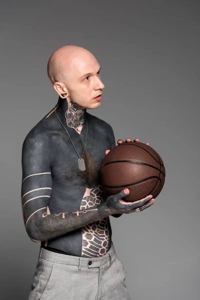 Uomo tatuato calvo senza maglietta che tiene palla da basket e distoglie lo sguardo isolato sul grigio — Foto stock