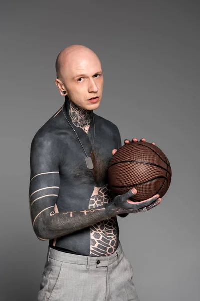 Bare-chested man with tattoos holding basketball ball and looking at camera isolated on grey — Stock Photo