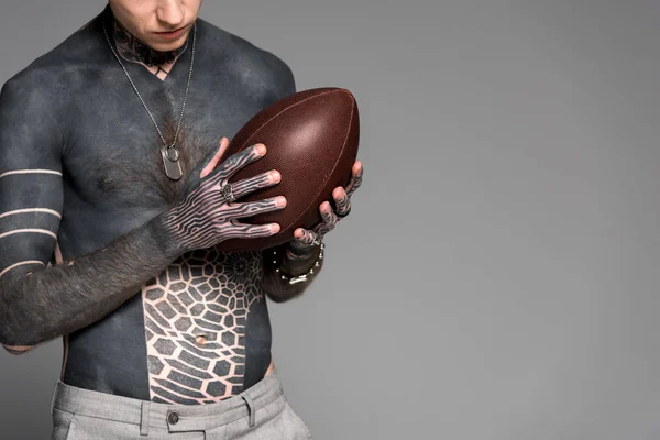 Plan recadré d'un homme à poitrine nue avec des tatouages tenant une balle de rugby isolée sur du gris — Photo de stock
