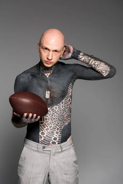 Лысый мужчина с татуировкой без рубашки, держащий мяч для регби и смотрящий на камеру, изолированную на сером — стоковое фото