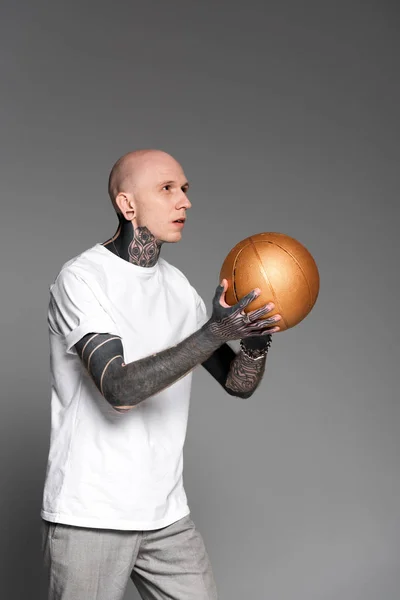 Tätowierter Mann mit Glatze in weißem T-Shirt spielt Basketball mit goldenem Ball isoliert auf grau — Stockfoto