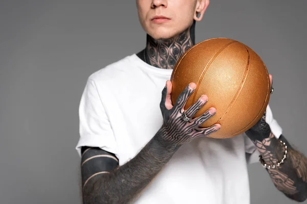 Tiro recortado de hombre tatuado en camiseta blanca sosteniendo pelota de baloncesto de oro aislado en gris - foto de stock