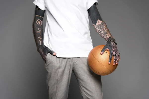 Colpo ritagliato di uomo tatuato in t-shirt bianca in piedi con pallone da basket dorato isolato su grigio — Foto stock