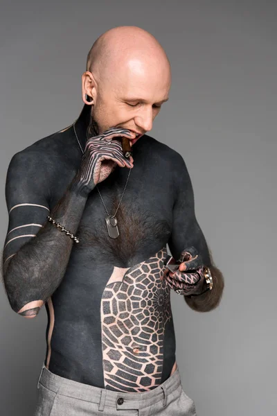 Улыбающийся мужчина без рубашки с татуировкой, держащий зажигалку и дымящуюся сигару изолированную на сером — стоковое фото