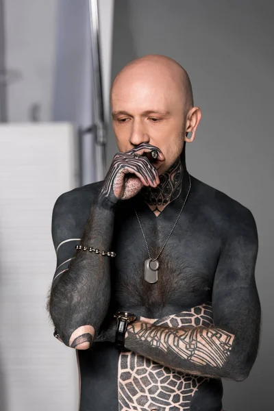 Bald shirtless man with tattoos smoking cigar and looking down on grey — Stock Photo