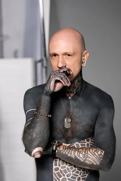 Bald shirtless man with tattoos smoking cigar and looking at camera on grey — Stock Photo