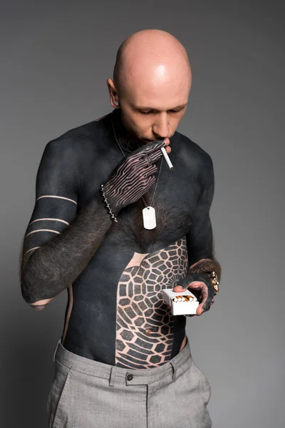 Careca sem camisa tatuado homem segurando caixa e fumar cigarro isolado no cinza — Fotografia de Stock