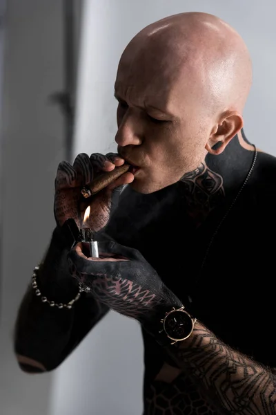 Bald shirtless tattooed man holding lighter and smoking cigar on grey — Stock Photo