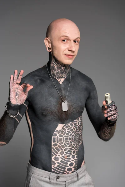 Hombre tatuado sin camisa con billetes de dólar que muestran signo de ok y sonriendo a la cámara aislado en gris - foto de stock