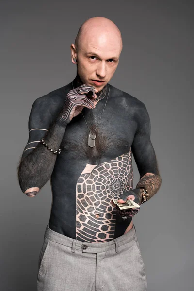 Hombre calvo sin camisa tatuado con cigarros sosteniendo billetes de dólar y mirando a la cámara aislada en gris - foto de stock