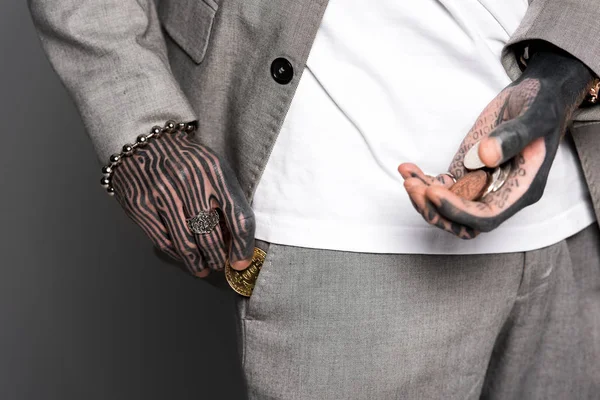 Cropped shot of tattooed man in suit holding coins and putting bitcoin in pocket isolated on grey — Stock Photo