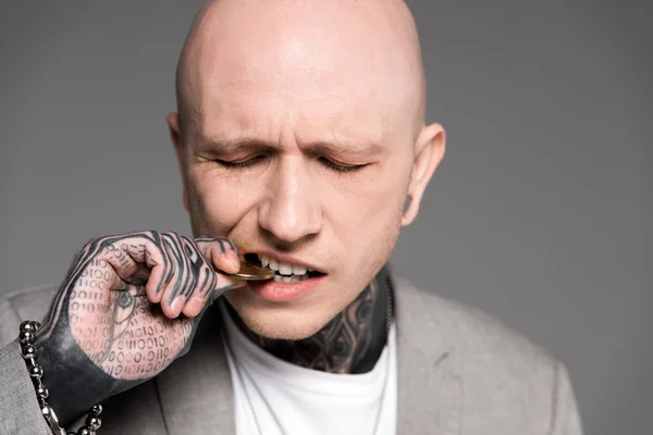 Close-up view of bald tattooed man biting bitcoin isolated on grey — Stock Photo