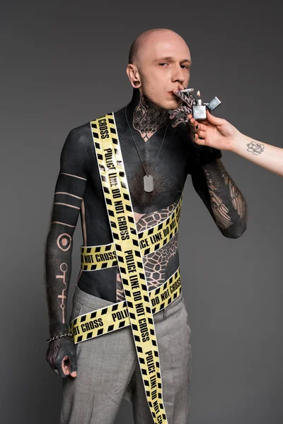 Cropped shot of someone holding lighter and tattooed man with police line around body smoking roll-up cigarette isolated on grey — Stock Photo