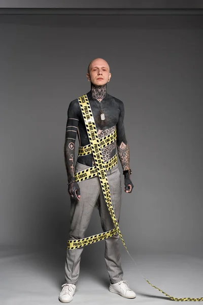Повнометражний вид на лисий татуйований чоловік з поліцейською лінією навколо тіла, дивлячись на камеру на сірому — стокове фото