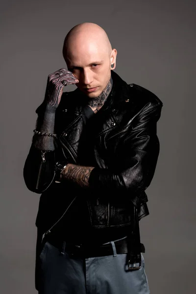 Confiado hombre tatuado en chaqueta de cuero de pie con la mano en la cara y mirando a la cámara aislada en gris - foto de stock