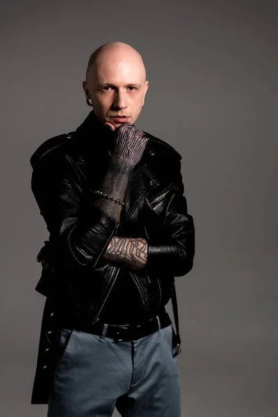 Stylish tattooed man in leather jacket standing with hand on chin and looking at camera isolated on grey — Stock Photo