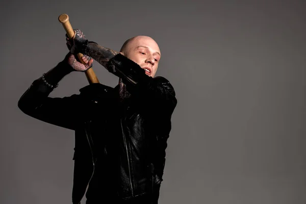 Aggressive bald tattooed man in leather jacket hitting with baseball bat isolated on grey — Stock Photo