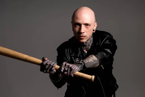 Agresivo hombre calvo tatuado en chaqueta de cuero sosteniendo bate de béisbol y mirando a la cámara aislada en gris - foto de stock