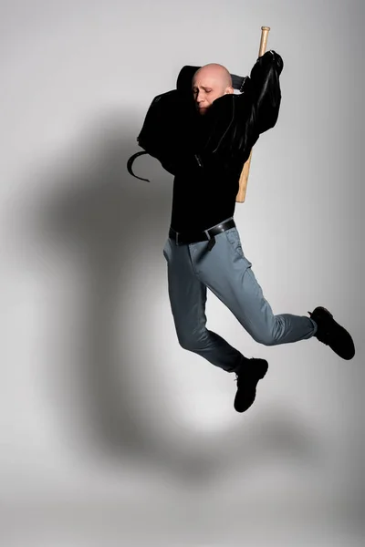 Homem careca agressivo em jaqueta de couro segurando bastão de beisebol e pulando em cinza — Fotografia de Stock