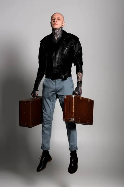 Full length view of handsome tattooed man in leather jacket holding suitcases and jumping on grey — Stock Photo