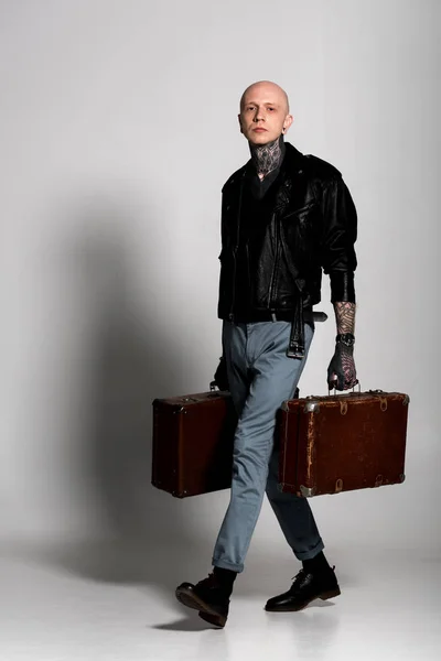 Hombre tatuado guapo en chaqueta de cuero sosteniendo maletas y mirando a la cámara en gris - foto de stock
