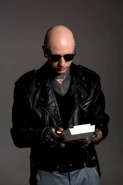 Лысый татуированный мужчина в кожаной куртке и солнцезащитных очках, держащий коробку с вкусным бургером, изолированным на сером — стоковое фото
