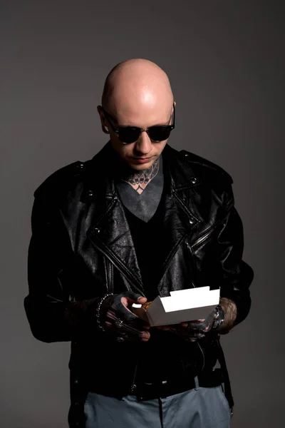 Bald tattooed man in leather jacket and sunglasses holding box with tasty burger isolated on grey — Stock Photo