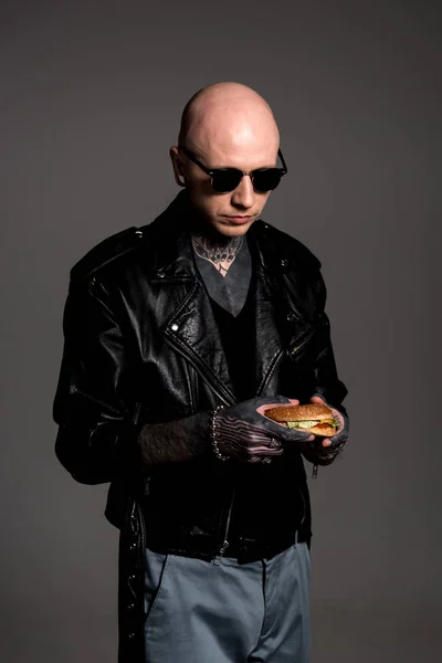 Elegante uomo pelato tatuato in giacca di pelle e occhiali da sole guardando delizioso hamburger in mani isolate su grigio — Foto stock