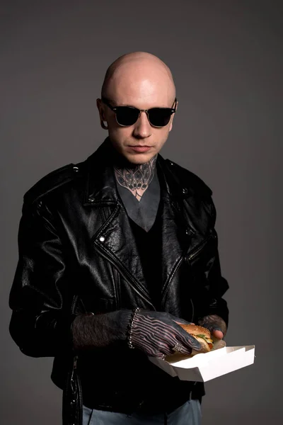 Elegante uomo tatuato in giacca di pelle e occhiali da sole scatola di tenuta con hamburger isolato su grigio — Foto stock